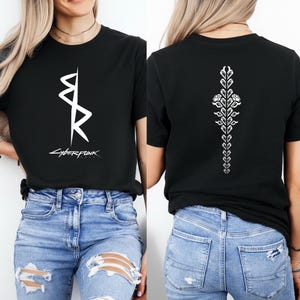 May include: Black t-shirt with a white graphic on the front and back. The front graphic is a stylized design with the word "CYBERPUNK" below it. The back features a vertical floral design. The shirt is paired with blue jeans.