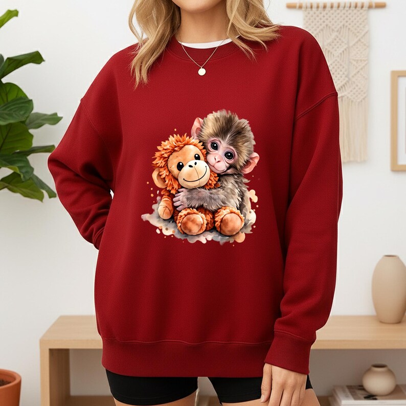 May include: A maroon sweatshirt featuring a graphic of two hugging monkeys. One monkey is brown and the other is orange. The sweatshirt has a crew neck and long sleeves. The image is a cartoon style.