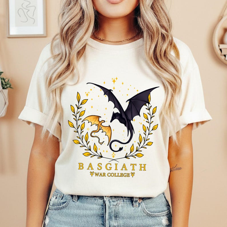May include: Cream-colored t-shirt featuring a graphic of two dragons, one black and one gold, within a gold leaf wreath. The text "BASGIATH WAR COLLEGE" is printed below the dragons in a serif font.