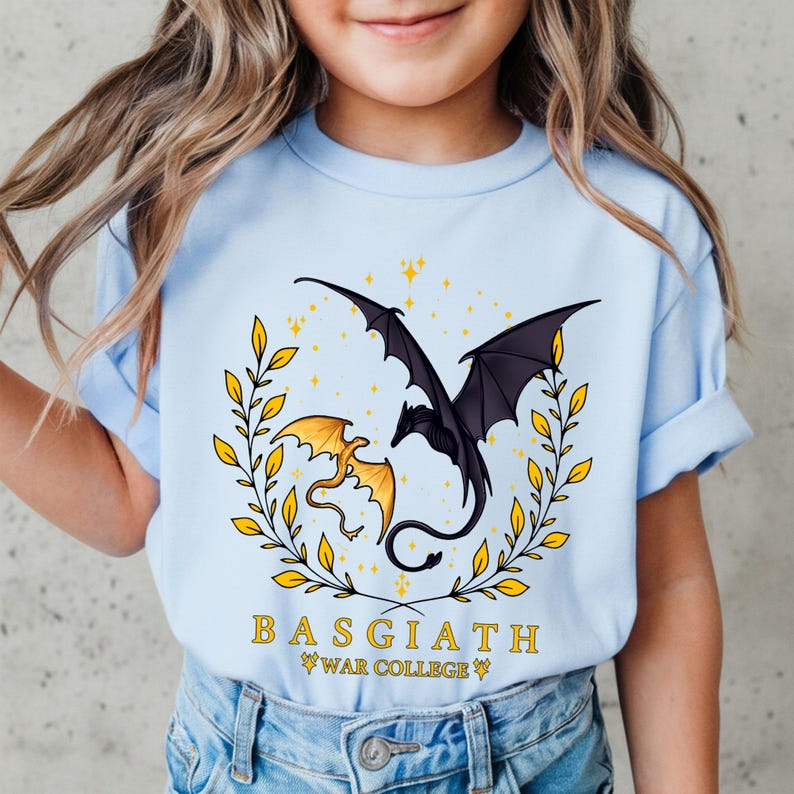 May include: Light blue t-shirt featuring a dragon design. The graphic includes two dragons, one gold and one black, surrounded by a gold wreath and stars. The text "BASGIATH WAR COLLEGE" is printed below the dragons.