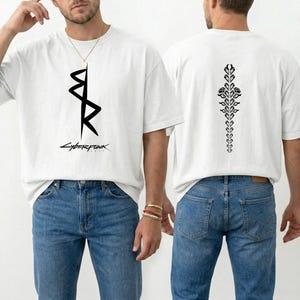 May include: White t-shirt with a black graphic on the front and back. The front features a stylized design with the word "Cyberpunk" below. The back has a vertical floral design. The shirt is worn with blue jeans.