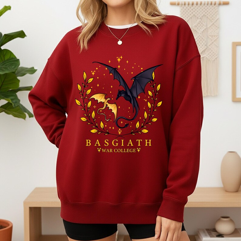 May include: A maroon sweatshirt featuring a dragon graphic. The design includes a black dragon with outstretched wings and a smaller gold dragon, surrounded by a gold leaf wreath and stars. The text "BASGIATH WAR COLLEGE" is printed below the dragons.