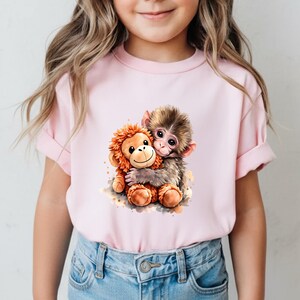 May include: A light pink t-shirt featuring a watercolor illustration of two hugging monkeys. One monkey is brown and the other is orange. The t-shirt has short sleeves and is made of a soft material.