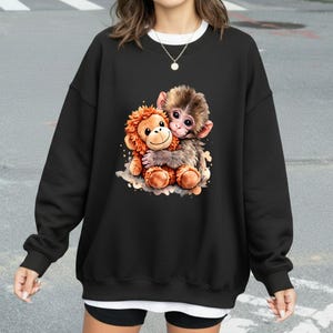 May include: Black sweatshirt featuring a colorful illustration of two hugging monkeys. One monkey is brown and the other is orange. The sweatshirt has a crew neck and long sleeves.