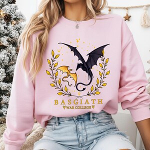 May include: Pink sweatshirt featuring a black and gold dragon design with the text "BASGIATH WAR COLLEGE". The graphic includes two dragons, one black and one gold, surrounded by a gold laurel wreath and stars.