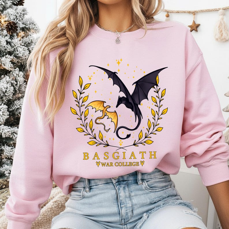 May include: Pink sweatshirt featuring a black and gold dragon design with the text "BASGIATH WAR COLLEGE". The graphic includes two dragons, one black and one gold, surrounded by a gold laurel wreath and stars.