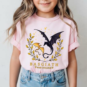 May include: A light pink t-shirt featuring a graphic of two dragons, one black and one gold, within a gold laurel wreath. The text "BASGIATH WAR COLLEGE" is printed below the graphic. The shirt is worn by a person.