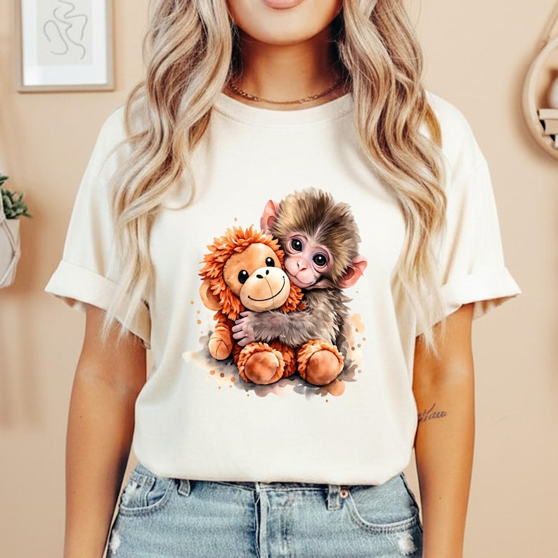 May include: Cream-colored t-shirt featuring a watercolor illustration of two hugging monkeys. One monkey is brown and gray, the other is orange and plush. The design is centered on the shirt, with a soft, watercolor effect.
