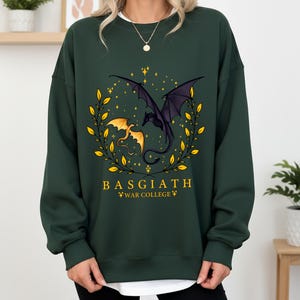 May include: Dark green sweatshirt featuring a dragon design. The graphic includes two dragons, one gold and one black, surrounded by a gold wreath and stars. The text "BASGIATH WAR COLLEGE" is printed below the dragons.