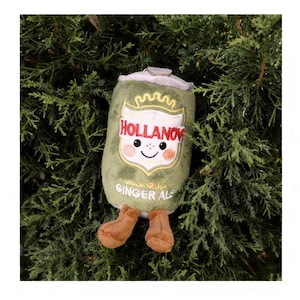 May include: A green plush toy in the shape of a ginger ale can, featuring the word "Hollanov" and a smiling face. The toy has brown feet and the words "Ginger Ale" are at the bottom. The background is green foliage.