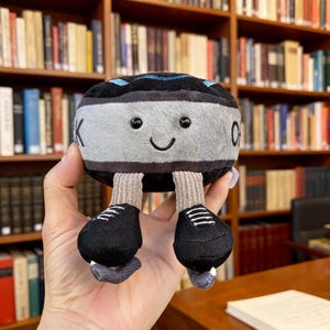 Op de afbeelding: Een knuffel in de vorm van een hockeypuck met een lachend gezicht, zwarte ogen en kleine benen met zwarte rolschaatsen. De puck is grijs en zwart met de letter "K" aan één kant. De achtergrond is een bibliotheek.