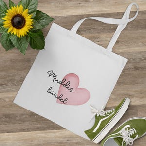 Könnte beinhalten: Weiße Tragetasche mit dem Text "Muddis büüdel" und einem rosa Herzdesign. Eine gelbe Sonnenblume und grüne Turnschuhe sind ebenfalls im Bild. Die Tasche hat zwei weiße Griffe.