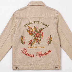 Puede incluir: Una chaqueta vaquera de color beige con un diseño bordado en la espalda. El bordado muestra un colibrí rodeado de motivos florales y de hojas en rojo, naranja y verde. El texto dice CHASE THE LIGHT AND LET YOUR Dreams Blossom.