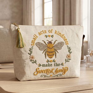 May include: A beige zippered pouch featuring an embroidered design of a honeybee centered within a floral wreath. The text reads, small acts of kindness make the sweetest honey. A green and gold tassel is attached to the zipper pull.