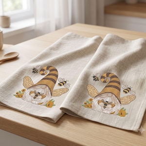 Puede incluir: Un par de paños de cocina de color beige claro con una ilustración repetida de un gnomo con sombrero a rayas, alas y barba, rodeado de pequeñas flores amarillas y abejas voladoras. El diseño está impreso en tonos amarillos, marrones y negros.