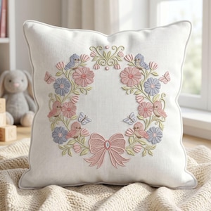 Könnte beinhalten: Quadratisches weißes Kissen mit einem gestickten Blumenkranz-Design. Der Kranz zeigt rosa und blaue Blumen, Vögel und Schmetterlinge mit einer rosa Schleife. Das Kissen hat einen hellgrauen Rand und liegt auf einer beigen Decke.