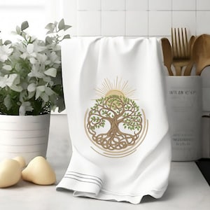 May include: White kitchen towel with a brown and green tree of life design with a sun above it. The tree is surrounded by a circle of braided rope.