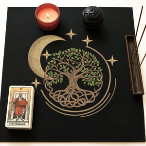 May include: A black cloth with a gold embroidered tree of life design, a crescent moon, and stars. There is a tarot card, a candle, incense sticks, and a black orb on the cloth.
