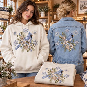 Pode incluir: Um moletom com capuz na cor creme e uma jaqueta jeans azul claro, ambos com um design bordado de um cardeal azul pousado entre galhos de pinheiro, pinhas, folhas de azevinho e flocos de neve azul claro na parte frontal.