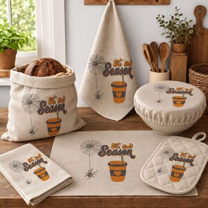 May include: A collection of beige kitchen textiles including a bread bag, a hanging towel, a bowl cover, a placemat, and a quilted potholder. Each item features a graphic of a spider web, a spider, a coffee cup, and the text Tis' the Season.