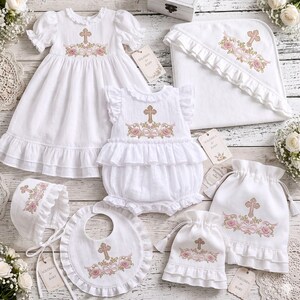 Può includere: Un set di abbigliamento bianco per neonati che include un vestito, una tutina con volant, un asciugamano con cappuccio, una cuffietta, una bavaglino e due sacchetti con coulisse. Ogni capo presenta un ricamo coordinato di una croce dorata.