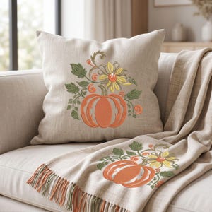 May include: A square beige throw cushion and a matching fringed throw blanket, both featuring an embroidered design of an orange pumpkin surrounded by green leaves, vines, and a yellow flower, creating a coordinated set for home decor.