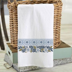 Peut inclure: Serviette blanche avec une bordure décorative ornée de motifs floraux et géométriques bleus et verts. La serviette est pliée et présentée devant un panier en osier, suggérant un textile de décoration intérieure ou de cuisine.