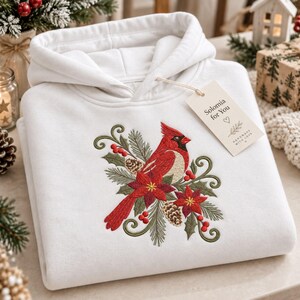May include: A white hooded sweatshirt featuring an embroidered design of a red cardinal bird perched among green pine needles, pinecones, red berries, and red poinsettia flowers. A beige rectangular tag attached to the hood reads Solomia for You.