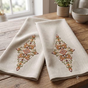May include: Two folded beige kitchen towels featuring an identical diamond-shaped embroidery pattern in shades of orange, green, and dark red. The intricate design consists of symmetrical floral and leaf motifs centered around a small square.
