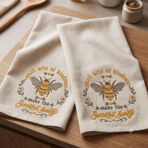May include: Two beige kitchen towels featuring embroidered designs of a honeybee surrounded by a floral wreath. The text reads, "small acts of kindness make the sweetest honey" in yellow and brown thread, centered on each fabric piece.