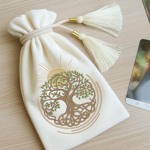 Puede incluir: Una bolsa de cordón de terciopelo blanco con borlas doradas. La bolsa presenta un diseño bordado de un árbol de la vida con raíces y ramas intrincadas, rodeado de un sol y círculos concéntricos.