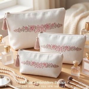 Könnte beinhalten: Set aus drei cremeweißen Kosmetiktaschen mit gestickten rosa Rosen und Schleifen. Jede Tasche hat einen goldenen Reißverschluss und einen passenden Quastenanhänger. Die Taschen sind auf einer Holzoberfläche mit Parfümflaschen und Make-up-Pinseln angeordnet.