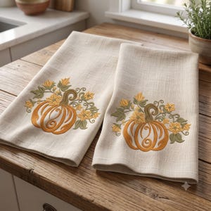 May include: Two beige kitchen towels featuring an embroidered design of an orange pumpkin surrounded by green leaves and yellow flowers. The fabric has a visible woven texture, and the embroidery is centered on the lower portion of each towel.