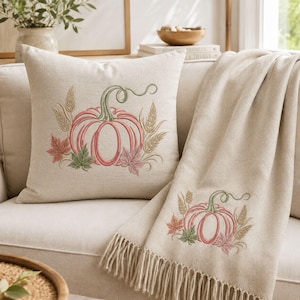 May include: A square throw cushion and a matching throw blanket in a light beige fabric. Both items feature an embroidered design of a pink pumpkin with a green stem, surrounded by green and pink maple leaves and stalks of wheat in a neutral tone.