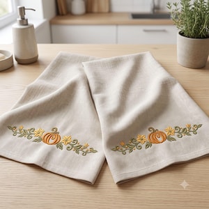 May include: Two light beige kitchen towels featuring an embroidered design of orange pumpkins, yellow flowers, and green vines. The embroidery is positioned horizontally across the lower section of each fabric towel, showcasing detailed stitching.
