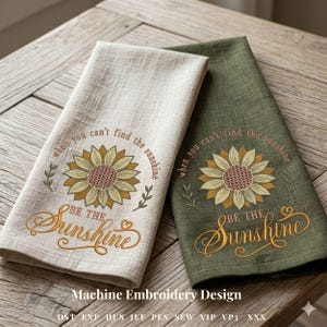 May include: Two embroidered kitchen towels, one beige and one olive green, with a sunflower design. The text reads "When you can't find the sunshine, be the sunshine." The towels are folded and displayed on a wooden surface.