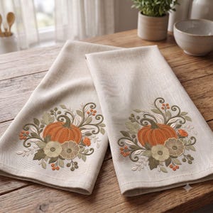 May include: Two beige kitchen towels featuring embroidered designs of orange pumpkins surrounded by sage green leaves, small flowers, and orange berries. The embroidery is centered on the lower portion of each folded fabric towel.