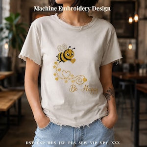 Puede incluir: Camiseta color crema con cuello y mangas desgastados. La camiseta presenta un diseño de bordado a máquina de una abeja de dibujos animados con un corazón, un rastro de corazones y estrellas, y las palabras "Be Happy" en hilo dorado.