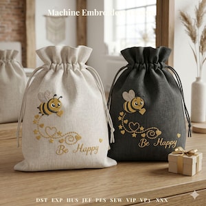 Puede incluir: Dos bolsas de regalo con cordón, una beige y otra gris antracita, con diseños bordados de abejas y corazones. El texto "Be Happy" está bordado debajo de la abeja. Las bolsas están sobre una superficie de madera.