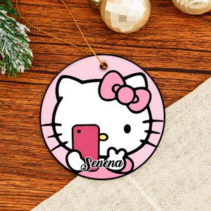 May include: A round pink and white ceramic ornament with a cartoon cat design. The cat is holding a pink phone and has a pink bow. The name "Serena" is written below. It has a gold hanging string.