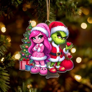 May include: A Christmas ornament featuring cartoon characters in festive attire. A pink-haired character in a pink outfit and a green-skinned character in a Santa suit stand together. A Christmas tree and gifts are in the background. The name "Serena" is at the bottom.