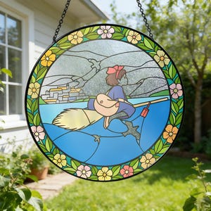 Puede incluir: Ventana redonda de vidrieras con una ni&ntilde;a de dibujos animados en una escoba sobre el agua. El dise&ntilde;o incluye una ciudad costera, monta&ntilde;as y un borde floral con acentos amarillos, rosas y verdes. La ventana est&aacute; enmarcada en negro y cuelga de una cadena.