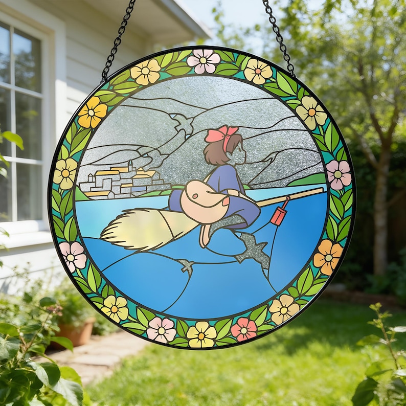 Puede incluir: Ventana redonda de vidrieras con una ni&ntilde;a de dibujos animados en una escoba sobre el agua. El dise&ntilde;o incluye una ciudad costera, monta&ntilde;as y un borde floral con acentos amarillos, rosas y verdes. La ventana est&aacute; enmarcada en negro y cuelga de una cadena.