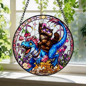 May include: A circular stained glass suncatcher featuring a brown cat wearing purple sunglasses and a crown, riding a blue dinosaur. The scene includes pink flowers, a yellow brick path, and a black metal chain for hanging against a window.