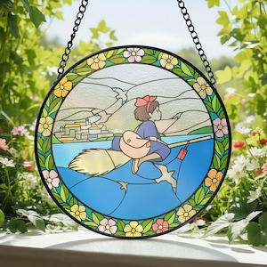 Puede incluir: Vidriera redonda que representa a una ni&ntilde;a de dibujos animados montada en una escoba sobre agua azul. La obra de arte tiene un marco de metal negro y una cadena para colgar. El borde est&aacute; decorado con flores coloridas y hojas verdes. Una pieza encantadora para cualquier habitaci&oacute;n.