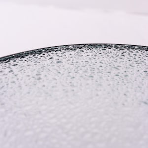 Puede incluir: Primer plano de un plato de vidrio transparente con una superficie texturizada. El plato tiene un borde oscuro y un patr&oacute;n de peque&ntilde;as formas irregulares en su superficie. El vidrio parece estar ligeramente te&ntilde;ido, con un sutil tono azul-verdoso.