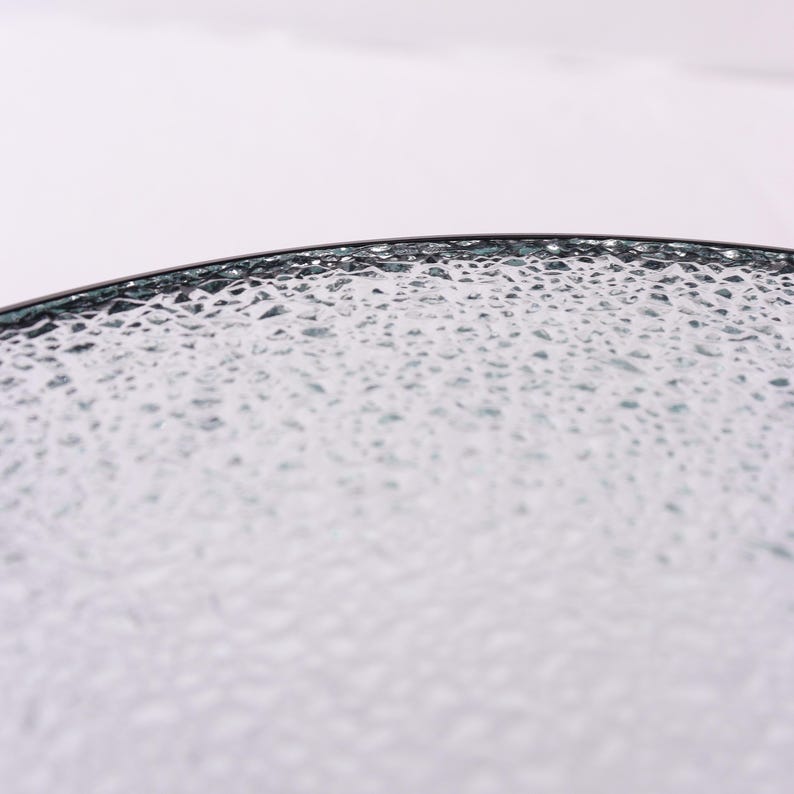 Puede incluir: Primer plano de un plato de vidrio transparente con una superficie texturizada. El plato tiene un borde oscuro y un patr&oacute;n de peque&ntilde;as formas irregulares en su superficie. El vidrio parece estar ligeramente te&ntilde;ido, con un sutil tono azul-verdoso.
