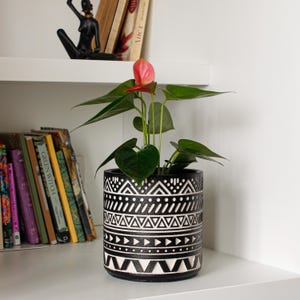 Op de afbeelding: Een roze Anthurium plant in een zwarte en witte geometrische gepatroneerde keramische pot. De pot heeft een witte achtergrond met zwarte geometrische vormen.