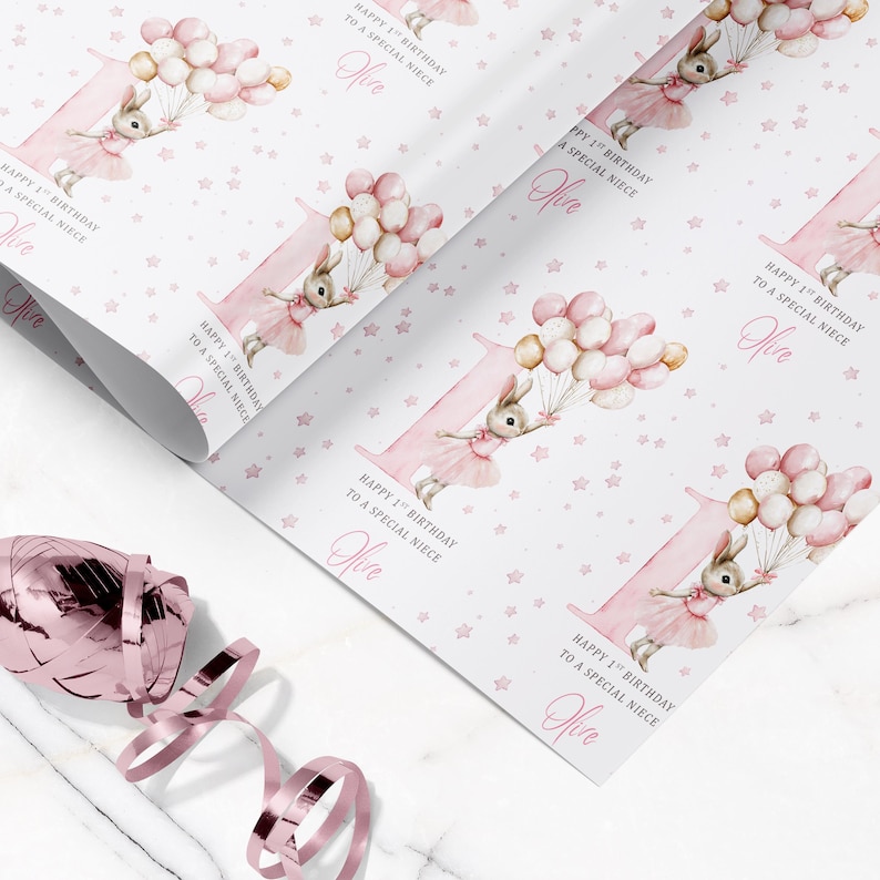 May include: Pink wrapping paper featuring a large number 1 with a bunny holding balloons. The paper has a repeating pattern of stars and the text "Happy 1st Birthday to a special niece" and the name "Olive".