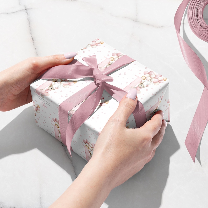 May include: A wrapped gift box with a pink satin ribbon tied in a bow. The gift wrap is white with a pattern of pink flowers and bunnies. The ribbon is a solid pink color.